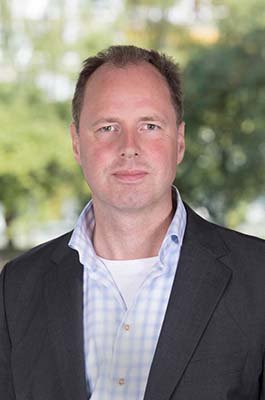 Portretfoto van advocaat Jan Paul van der Kooij, partner bij Van der Kooij Besters Advocaten, gefotografeerd vanaf de borst omhoog tegen een zachte, onscherpe groene achtergrond die lijkt op bomen of een parkachtige omgeving. Hij staat recht naar de camera toe en kijkt direct vooruit met een rustige, zelfverzekerde en toegankelijke uitstraling. Hij draagt een donker colbert over een lichtblauw geruit overhemd met open kraag, wat een nette maar toegankelijke professionele uitstraling geeft die past bij een advocaat die cliënten persoonlijk begeleidt. De foto illustreert het profiel van Jan Paul van der Kooij als ervaren advocaat in letselschade en personen- en familierecht, die al meer dan 25 jaar mensen bijstaat bij ingrijpende gebeurtenissen.